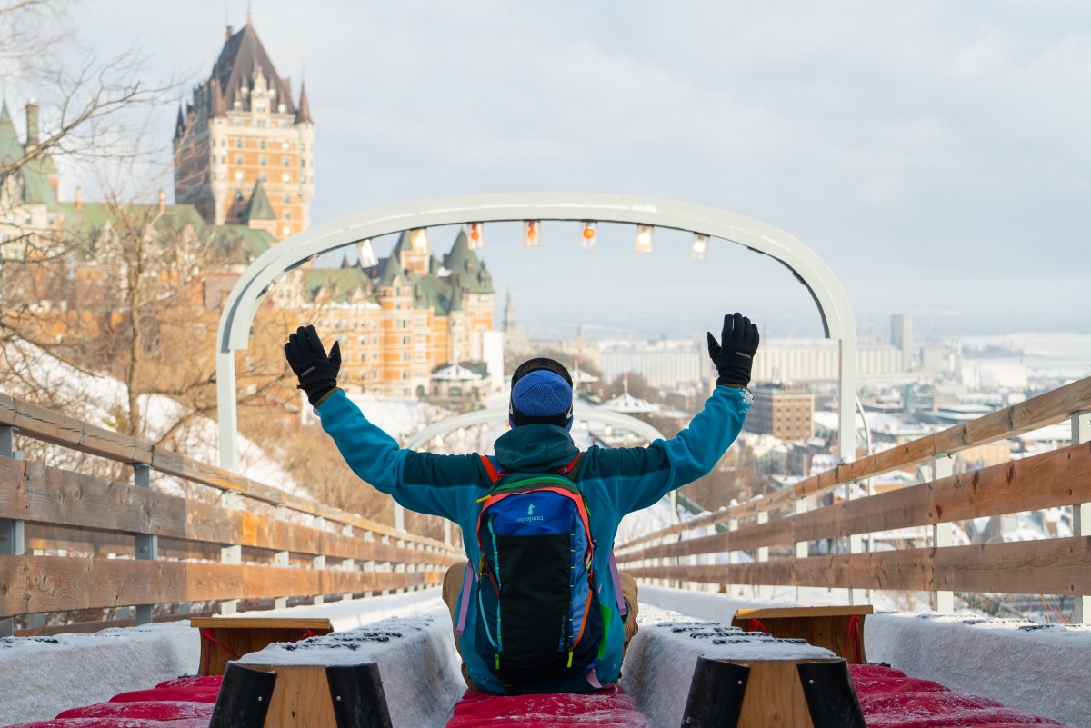 Person with backpack waving on a snowy slide, large building in background.