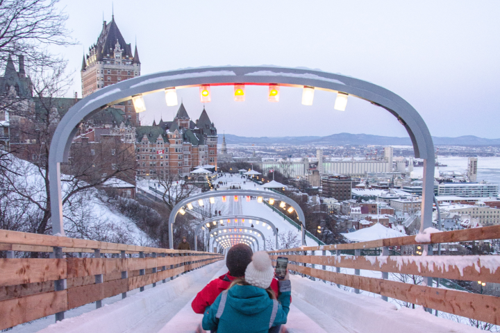 Two people sledding on an icy track with a castle-like building and city in the background.