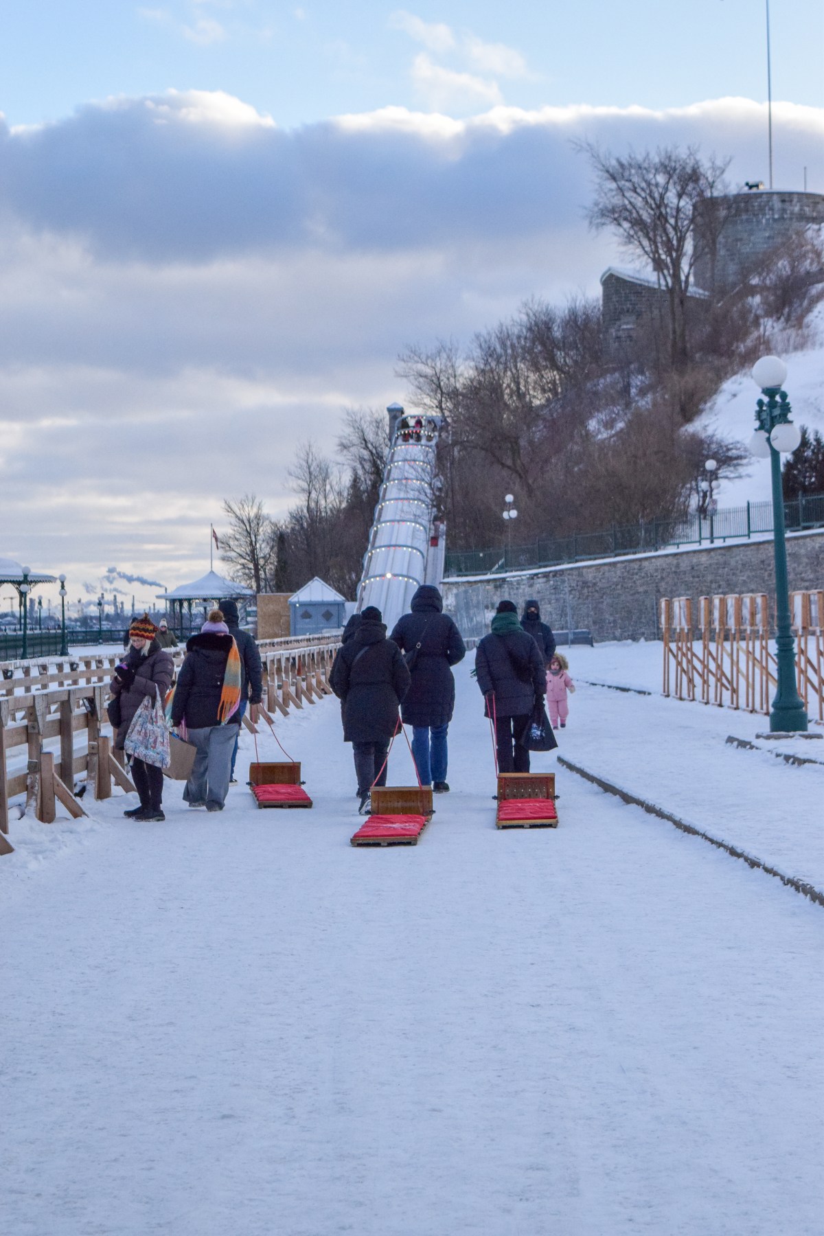 People pulling sleds on a snowy path towards a tall ice slide with cloudy sky.