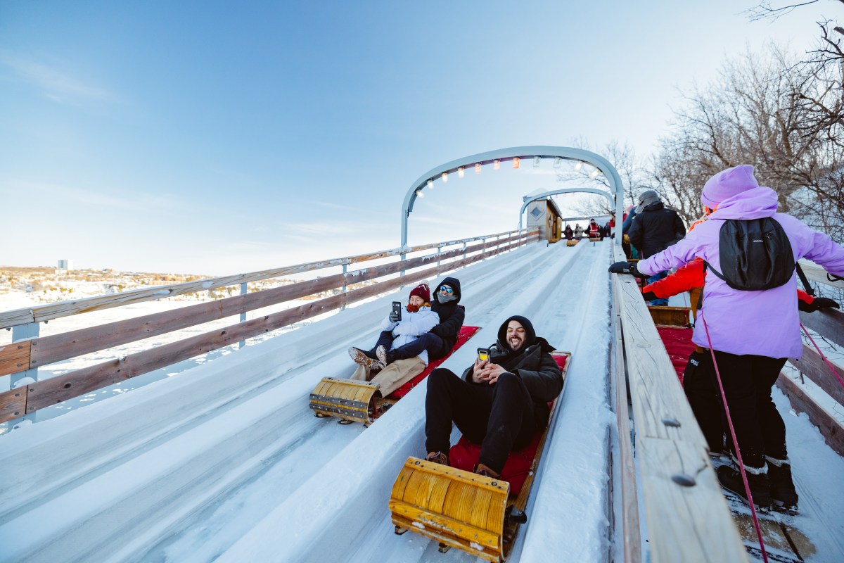 People sledding on a snowy slide with clear blue sky and trees in the background.
