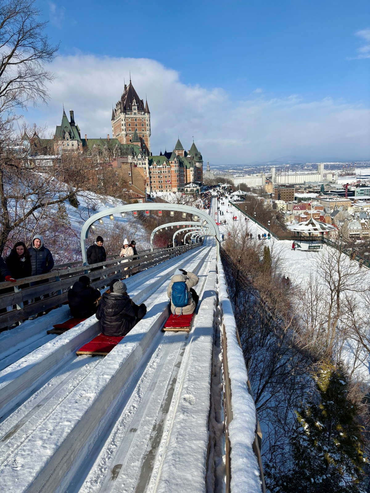 People sledding on snowy track beside historic castle with city view.