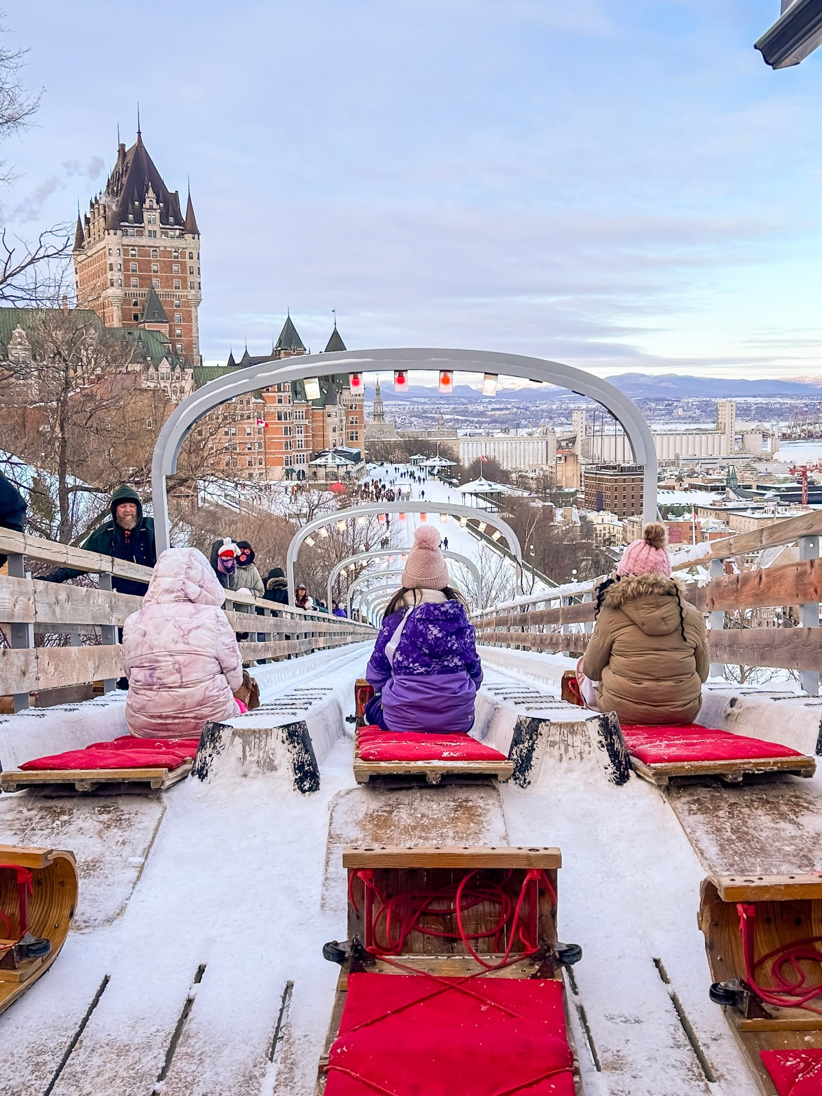 People sledding down a snowy slide with a castle in the background and scenic city view.