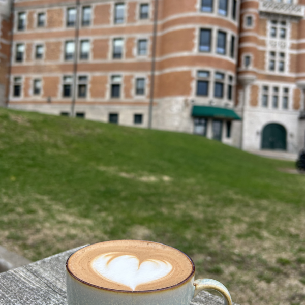 Cup of coffee with heart latte art on outdoor table, building in background.