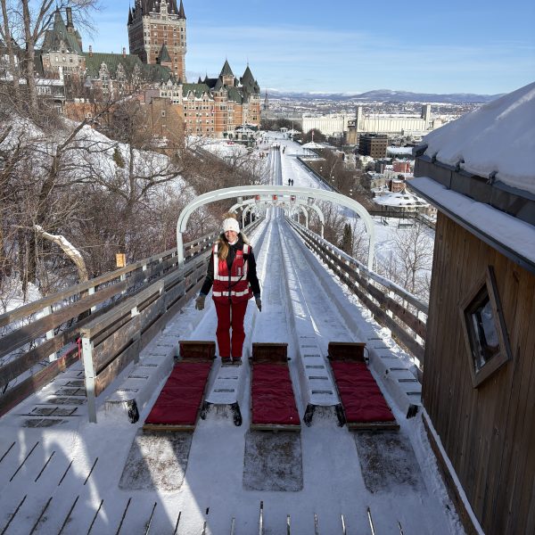 Person in red suit stands by sleds on a snowy slide with city buildings in background.