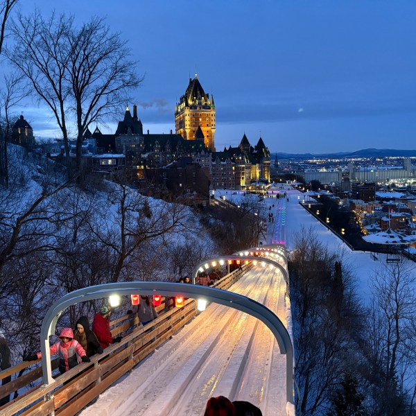 Snowy sledding track with people, overlooking a lit castle and city at dusk.